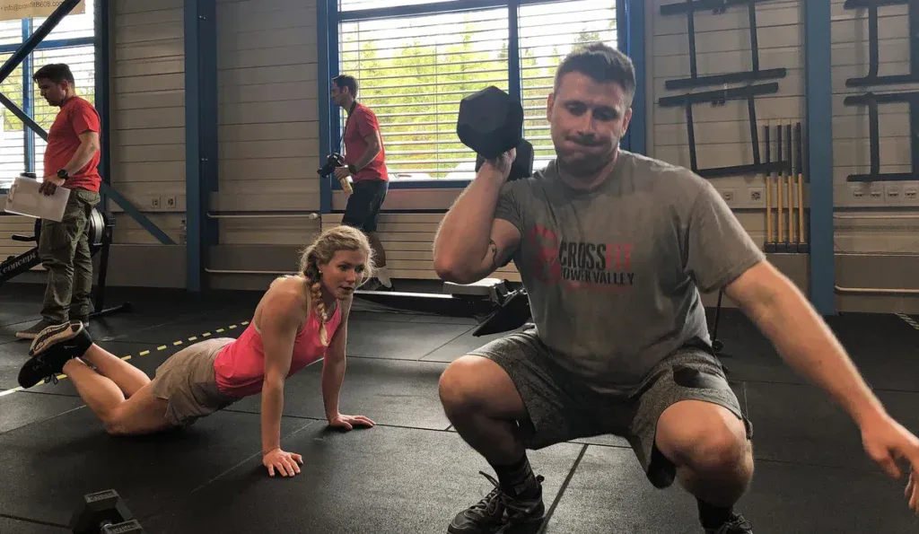 Zawody CrossFit: zawodnik z hantlem, obok zawodniczka robi burpees, sędzia z kartą wyników w tle